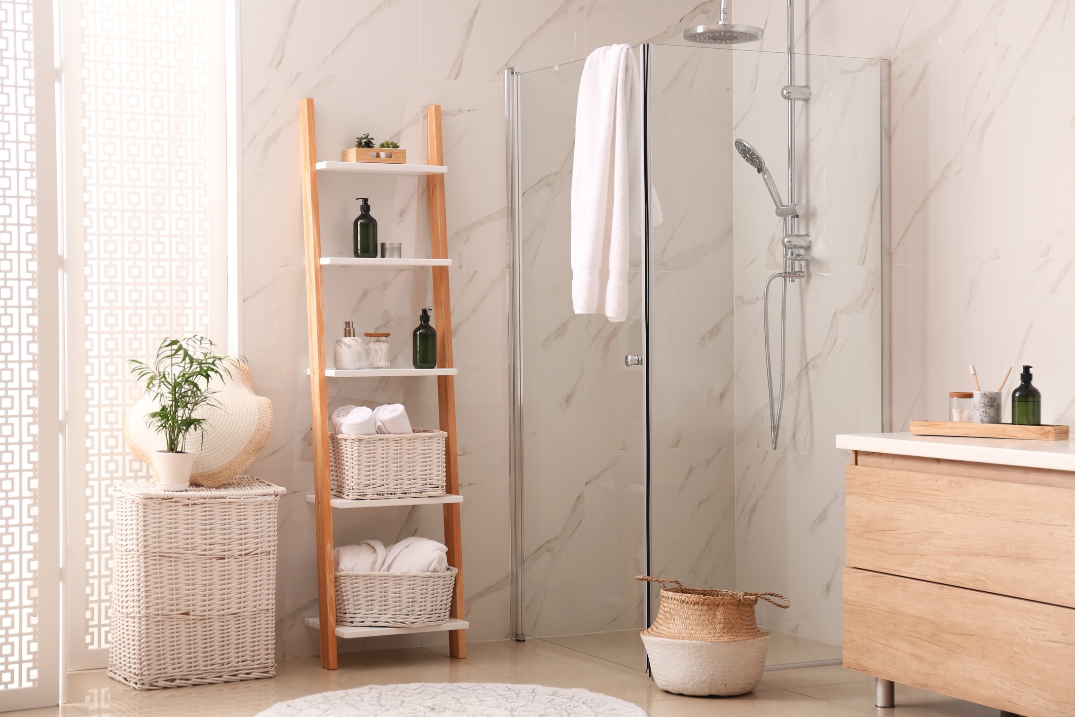 Modern bathroom with a glass shower, wood shelf holding towels and toiletries, wicker baskets, a small potted plant, and a wooden vanity with a countertop. Cream-colored walls and a decorative privacy screen add to the serene ambiance.