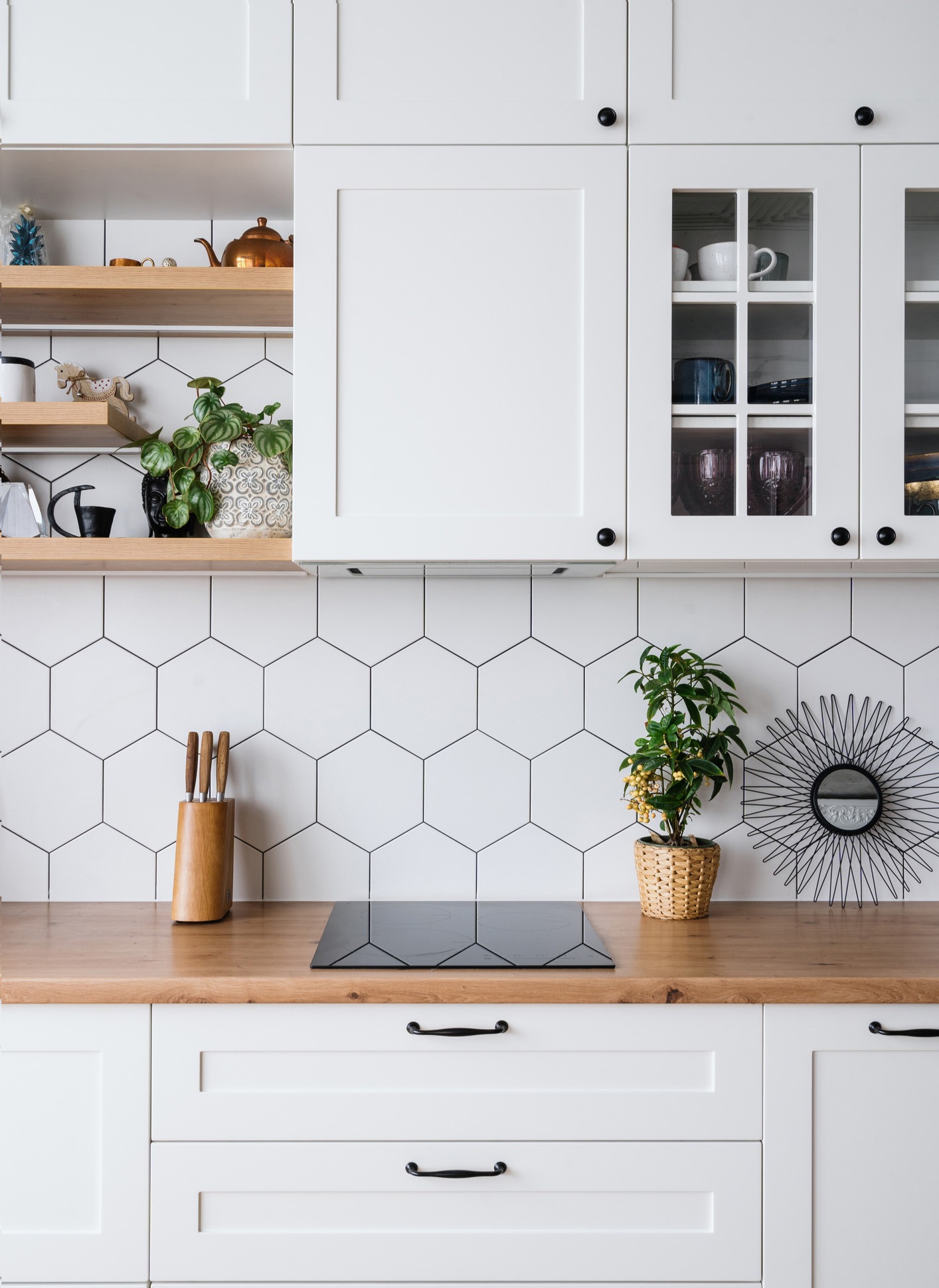 A modern kitchen with white cabinets, hexagonal tile backsplash, and wooden countertops. Shelves display plants, a teapot, and cups. A small plant sits near a decorative black wire wall piece above a stove.