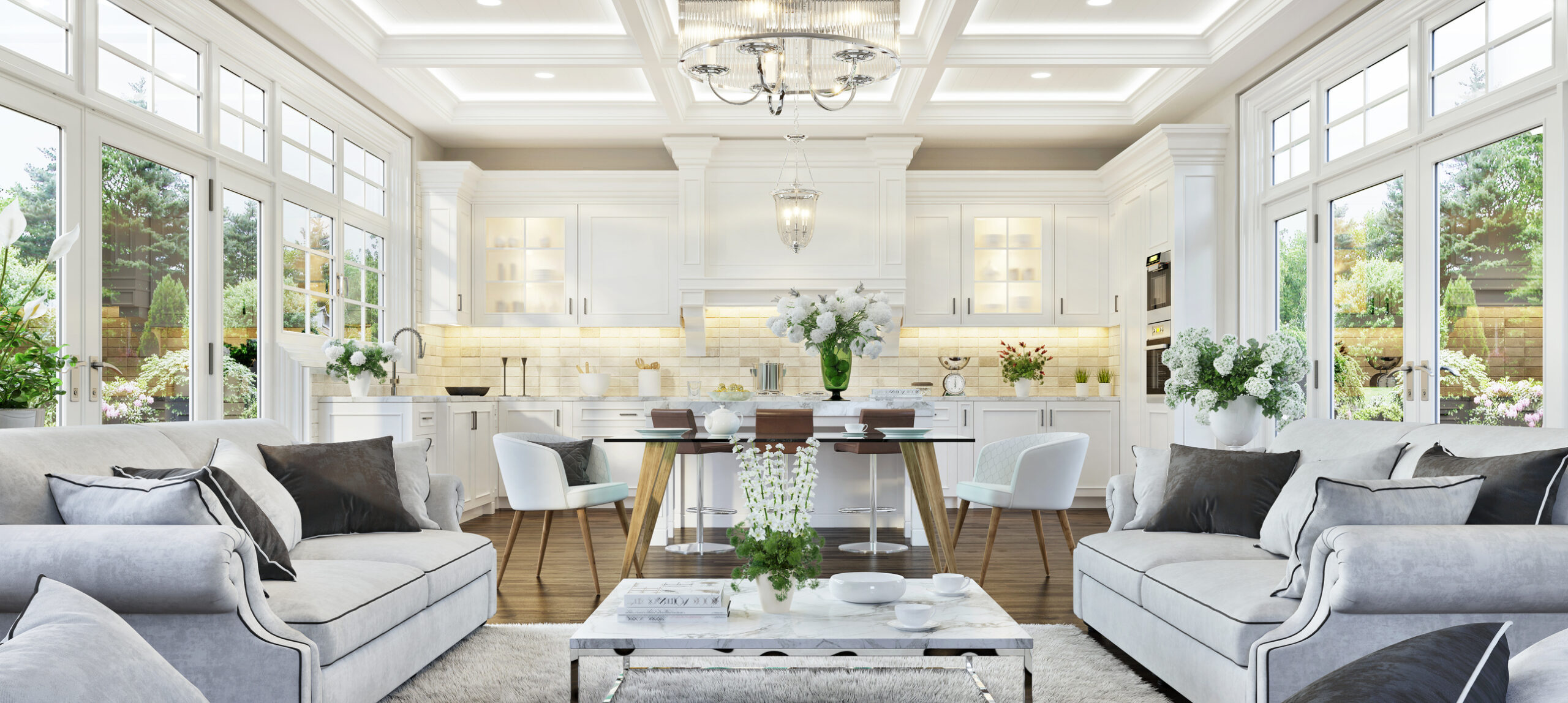 A spacious, elegant living room and kitchen with white decor. Large windows provide natural light, highlighting two gray sofas, a glass coffee table, and a dining table with white chairs. Lush greenery is visible outside.