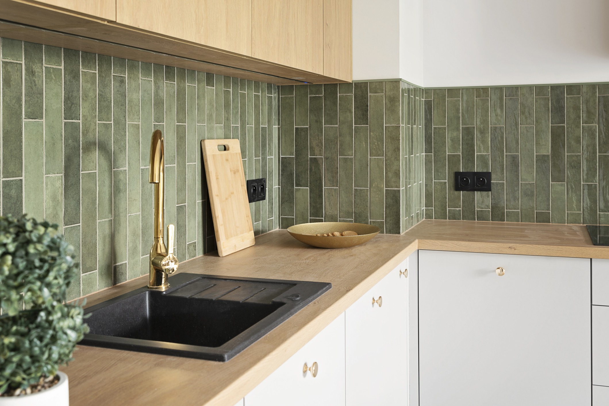 Modern kitchen with green tiled backsplash, light wooden countertops, white cabinets, and a black sink. A gold faucet and a wooden cutting board are near the sink. A potted plant and a bowl with fruit decorate the counter.